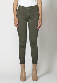 Pantalones ajustados de color verde oliva con intrincados bordados cerca de los tobillos, combinados con botas marrones beige y una camiseta blanca.