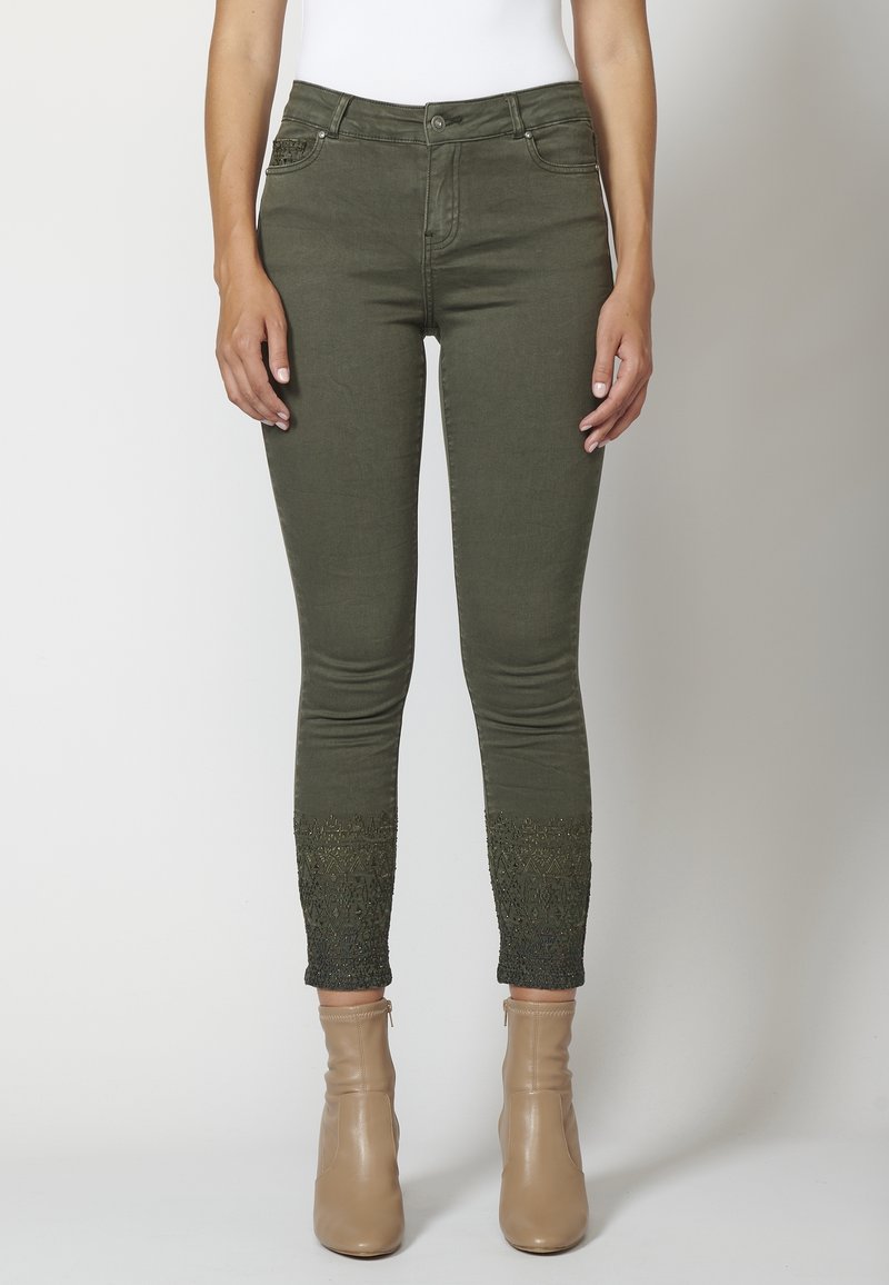 Pantalones ajustados de color verde oliva con intrincados bordados cerca de los tobillos, combinados con botas marrones beige y una camiseta blanca.