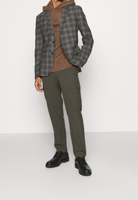 Blazer de cuadros gris, sudadera con capucha marrón y pantalones cargo verde oliva. Zapatillas negras con acabado texturizado. Look casual en capas.
