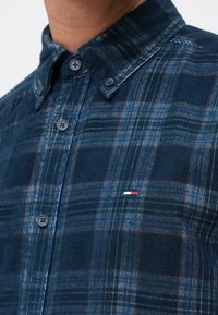 Camisa de cuadros azul con cuello botón, que presenta cuadros de color azul oscuro, azul claro y burdeos. Pequeño logo bordado en rojo, blanco y azul.