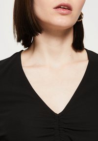 Schwarzes V-Ausschnitt-Top mit gerafftem Stoff in der Mitte, glatter Textur, figurbetontem Schnitt und kurzen Ärmeln. Dezenter Glanz im Material sichtbar.