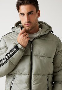 Chaqueta acolchada verde claro con capucha, con un diseño de acolchado texturizado. Branding negro "GUESS" en la manga y bolsillos con cremallera en el frente.