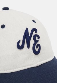 Cappello bianco con accenti blu scuro, caratterizzato da un design ricamato "NE". Tessuto di cotone con una texture liscia, forma strutturata e sei fori di ventilazione.