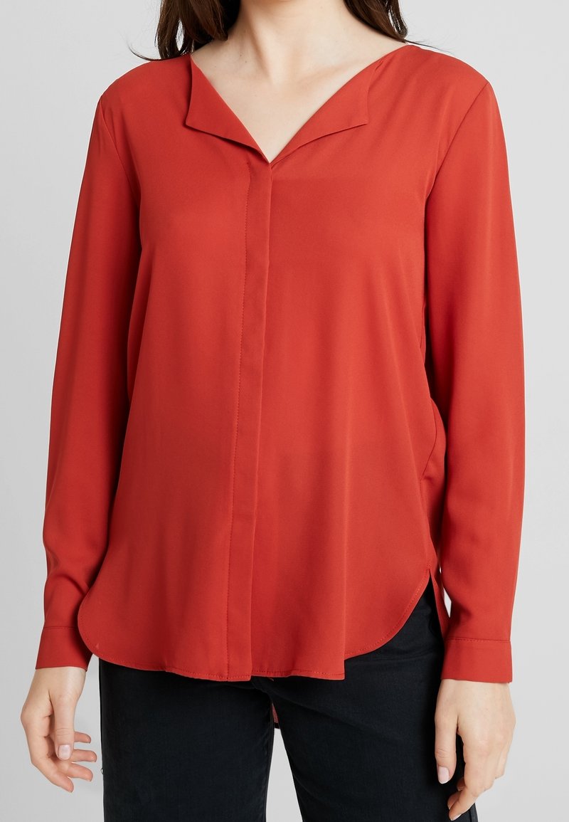 Blouse rouge à manches longues avec un décolleté en V, fabriquée en tissu lisse. Présente un ourlet incurvé et des détails de couture subtils. Portée avec un pantalon sombre.