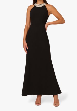 Kobieta ubrana w czarną, długą do ziemi suknię bez rękawów z dekoltem ozdobionym perłami, stojąca z jedną ręką na biodrze, w beżowych, otwartych na palce butach na obcasie.
