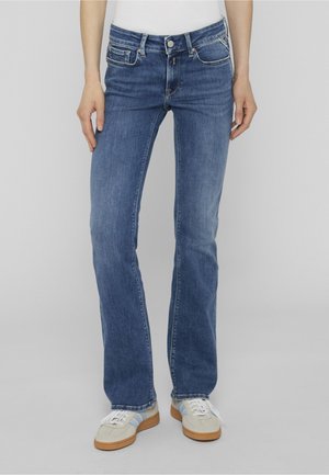 Person trägt blaue Jeans mit geradem Bein und mittlerer Leibhöhe sowie beige Sneaker mit weißen Schnürsenkeln vor schlichtem grauem Hintergrund.