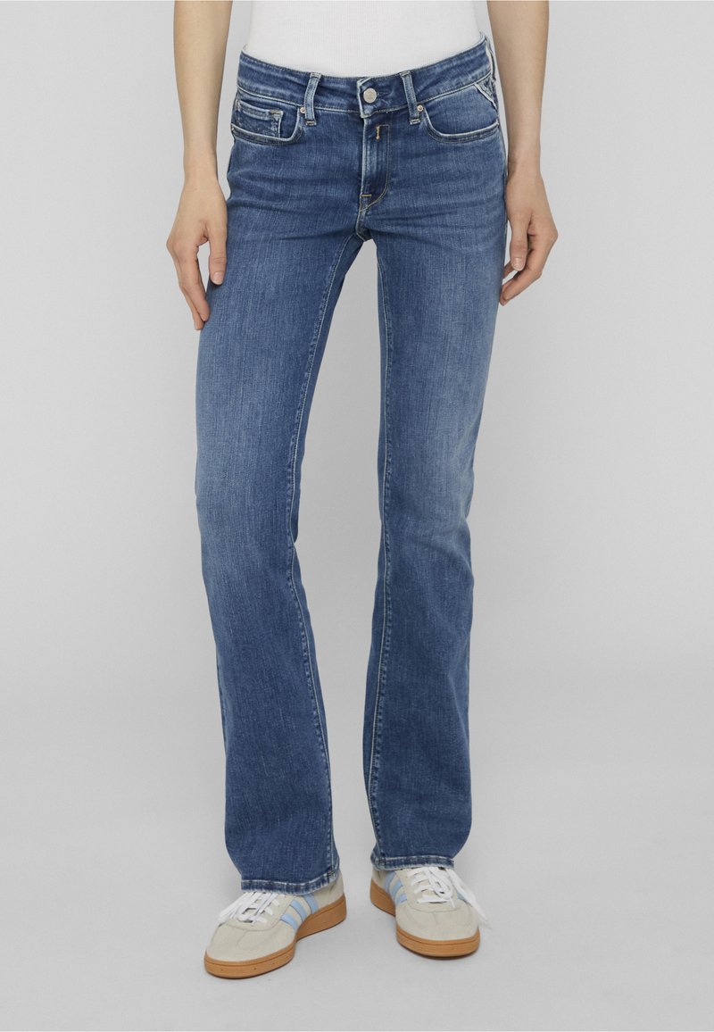 Personne portant un jean bleu taille mi-haute, coupe droite, et des baskets beiges avec des lacets blancs sur un fond gris uni.