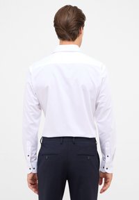 Witte langwerpige overhemd met knoopmanchetten, met een gladde textuur. Gedragen met donkerblauwe broek, met details aan de achterkant.