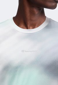 Άνδρας με λευκό T-shirt με στρογγυλή λαιμόκοψη και λεπτό λογότυπο Champion στο κέντρο του στήθους, με το κεφάλι ελαφρώς γυρισμένο προς τα δεξιά.