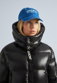 Donna in un giubbotto a collo alto nero e cappellino blu con logo "JACKIT Ski & Social Club", che guarda di lato.