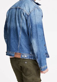 Denim takki keskisinisessä sävyssä, jossa on terävä kaulus, nepparit ja ruskea nahkabrändilogo takana. Takissa on rento malli.