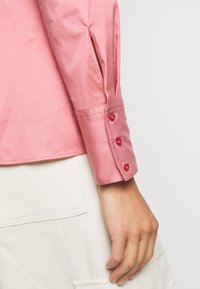 Camicia rosa in satin con polsini abbottonati, dotata di tre bottoni rossi e fessure ai polsi. Tessuto liscio e design su misura.