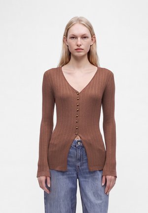 Junge Frau mit langen blonden Haaren trägt eine braune, gerippte, langärmelige, geknöpfte Strickjacke und lockere blaue Jeans vor einem schlichten hellen Hintergrund.