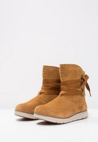 Tan kleuren suède enklaarzen met een zachte textuur, ronde neus, vetersluiting met accentstrikjes aan de zijkant en een crèmekleurige rubberen zool.