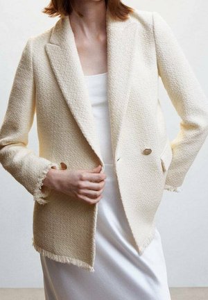 Mujer que lleva una blazer texturizada de color crema con botones dorados sobre una camiseta blanca sin mangas y una falda de satén blanca, ajustándose el frente de la blazer con la mano.