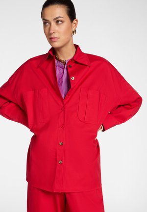 Femme portant une chemise rouge surdimensionnée boutonnée avec des poches avant par-dessus un haut violet, debout les mains sur les hanches devant un fond uni.