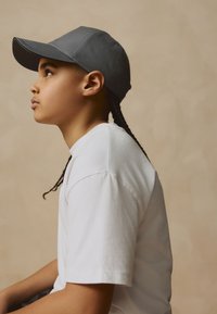 Graue Baseballkappe mit gebogenem Schirm, getragen zu einem einfachen weißen T-Shirt. Die Person hat sichtbares, glattes, geflochtenes Haar. Neutraler Hintergrund.