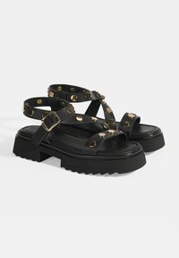 Black leather sandals with a chunky platform, featuring crossed straps, gold-colored studs, and a buckle detail. Textured sole for grip.