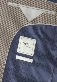 Veste à motif pied-de-poule marron et blanc avec doublure intérieure géométrique bleue et étiquette cousue "NEXT Tailoring".