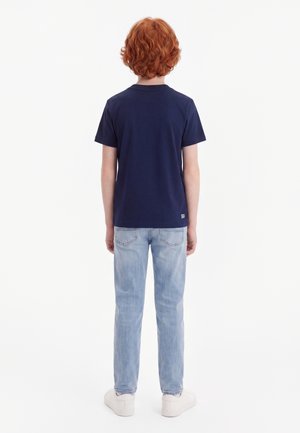 Niño con cabello rizado y rojo de espaldas, lleva puesta una camiseta azul marino, vaqueros azul claro y zapatillas blancas sobre un fondo blanco.