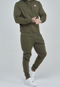 Olivgrüner Hoodie mit Fronttasche und Logo, kombiniert mit passenden Jogginghosen. Weiße Sneaker setzen einen Kontrast zum Outfit.