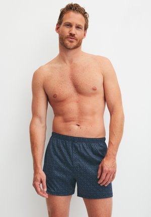 Man zonder shirt met krullend haar, gekleed in donkerblauwe korte broek met patroon, staand tegen een effen witte achtergrond.