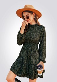 Abito verde con stampa leopardata, maniche lunghe, vita smock e orlo a balze a strati. Indossato con un cappello marrone e una tazza di caffè in mano.