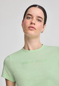 Camiseta de manga corta verde claro con cuello redondo. Presenta un logo texturizado de "POLO CLUB" en verde más oscuro, establecido en 2012. Tejido suave.