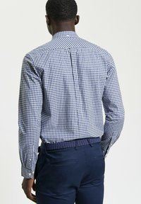 Camicia a scacchi blu scuro e bianca con colletto classico e polsini con bottoni, abbinata a pantaloni scuri e una cintura intrecciata, vista da dietro.