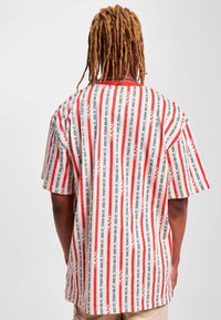 Camisa de manga corta y corte oversize en blanco con rayas verticales rojas y texto negro que se repite "SEE IT, THEN BE IT." Hecha de tela ligera.