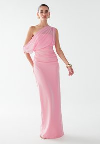 Jednoduché růžové šaty na jedno rameno s nařasenou texturou, splývavým chiffonovým přehozem, dlouhým střihem až na zem a přiléhavým siluetou. Zlacené náramkové šperky.