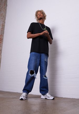 Sort T-shirt, slidset blå jeans med store huller, hvide sneakers og et sølvhalssmykke. Alt sammen foran en hvid murstensvæg.