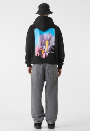 Svart hettegenser med et stort fargerikt grafisk design på ryggen som viser abstrakte blomsterdesigns. Grå baggy jeans og en svart bøttehatt kompletterer antrekket.