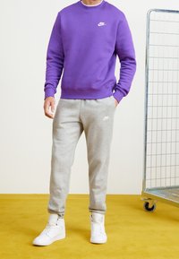 Lila fleece-tröja med rundad halsringning, kombinerad med ljusgrå träningsbyxor med en vit logotyp. Vita hög-top sneakers fulländar outfiten.