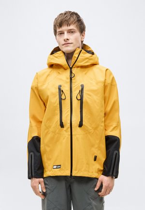 Jauns vīrietis valkā dzeltenu ūdensizturīgu jaku ar melnām rāvējslēdzēja detaļām un kapuci, stāv ar rokām atslābināti pie sāniem.