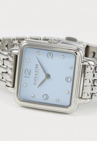Quadratische Stahl-Uhr mit einem hellblauen Zifferblatt, silbernen Ziffern und Kristallakzenten. Verbundenes Metallarmband mit glänzender Oberfläche.