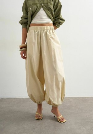 Persona che indossa pantaloni beige larghi con polsini elastici, un crop top bianco, una giacca verde oliva, sandali beige con tacco e braccialetti a un polso.