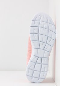 Semelle d'une basket rose à semelle en caoutchouc blanche appuyée contre un mur blanc sur une surface blanche.