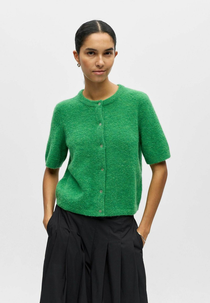 Femme aux cheveux foncés portant un pull vert à manches courtes boutonné et une jupe plissée noire, debout avec les mains dans les poches sur un fond blanc.