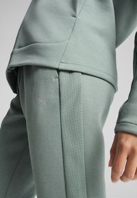 Un hoodie et un pantalon de sport verts en matière tricotée douce, avec des rayures texturées sur les côtés et un logo discret sur la jambe.