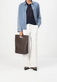 Personne portant une veste en denim clair, un haut en maille marine, un pantalon large blanc, des ballerines noires pointues, tenant un grand sac fourre-tout marron.