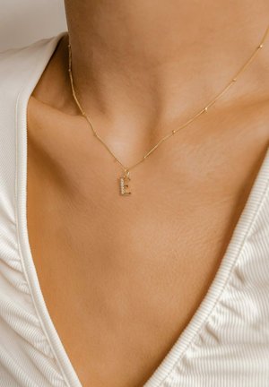 Goldene Halskette mit einem kleinen "E" Anhänger, verziert mit Strasssteinen, die auf nackter Haut aufliegt, vor einem weißen strukturierten Oberteil.