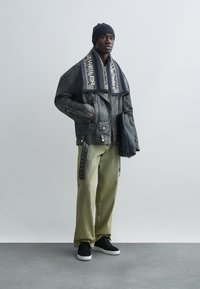 Svart läderjacka med silverdetaljer, kombinerad med löst sittande gröna byxor med ränder och svarta sneakers. Svart mössa och halsduk visas.