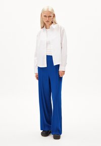 Witte blouse over een witte tanktop, gecombineerd met wijde blauwe broeken en zwarte schoenen. Minimalistisch ontwerp en levendig kleurcontrast.