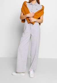 Combinaison en maille grise à jambes larges, taille ajustable avec cordon, associée à un pull moutarde porté sur les épaules et des baskets blanches.