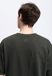 Donkergroen oversized t-shirt met een ronde halslijn, voorzien van een subtiel scharenlogo in een lichtere tint op de bovenrug. Gemaakt van katoen.