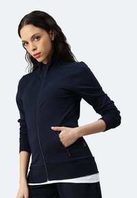 Giacca a zip blu navy in tessuto liscio con colletto alto, polsini aderenti e tasche laterali. Presenta un dettaglio di cucitura all'orlo.
