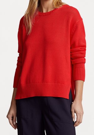 Kobieta ubrana w jaskrawoczerwony sweter z długim rękawem oraz ciemne, luźne spodnie, stojąca z ramionami swobodnie opuszczonymi wzdłuż boków.