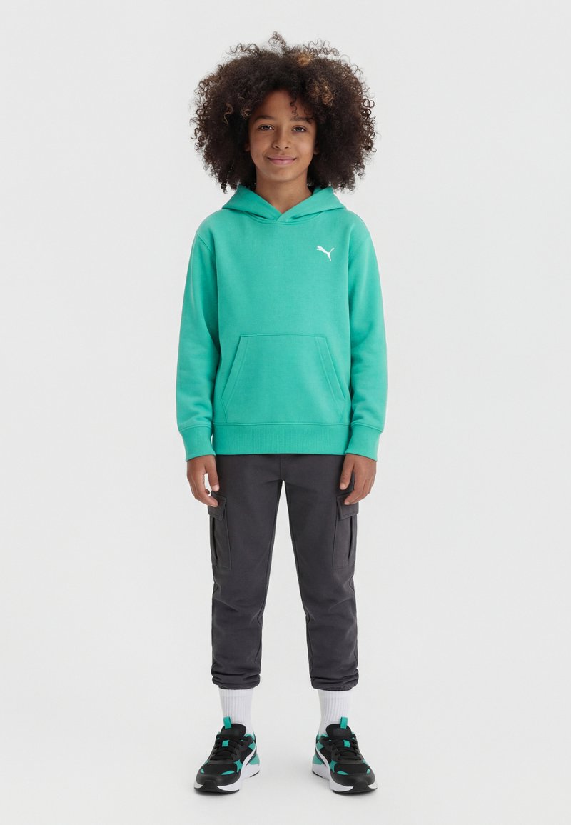 Παιδί με σγουρά μαλλιά που φοράει μια τιρκουάζ φούτερ Puma με κουκούλα, σκούρα γκρι cargo παντελόνια, λευκές κάλτσες και μαύρα-τιρκουάζ αθλητικά παπούτσια, στέκεται μπροστά σε λευκό φόντο.