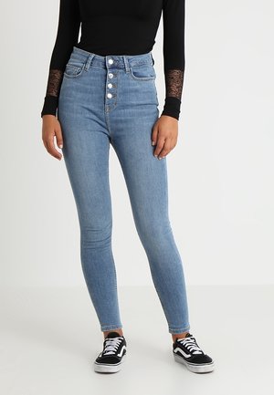 Personne portant un jean skinny taille haute bleu clair avec quatre boutons, un haut noir à manches longues avec des poignets en dentelle et des baskets noires et blanches.